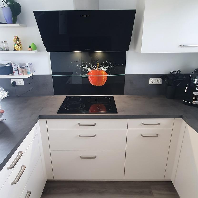 Küche nach Maß mit Glasrückwand, Kochfeld und Apfel-Motiv hinter Dunstabzugshaube.