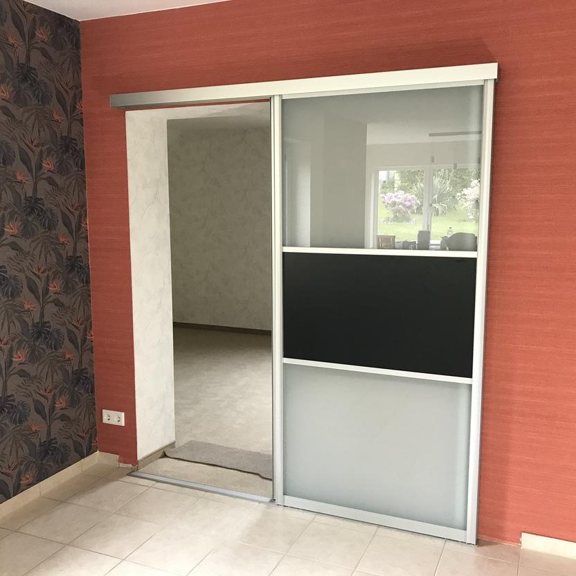 Moderne Schiebetür aus Glas mit schwarzem Mittelstreifen, montiert an roter Wand mit gemusterter Tapete und heller Fliesenboden.