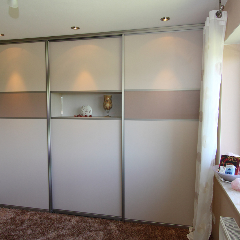 Großer Einbauschrank mit Schiebetüren in Beige und Glasakzent, beleuchtet, in einem hellen Schlafzimmer mit Teppichboden.