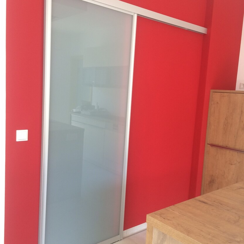Schlichte Schiebetür aus satiniertem Glas mit Aluminiumrahmen vor roter Wand, trennt moderne Küche und Essbereich.