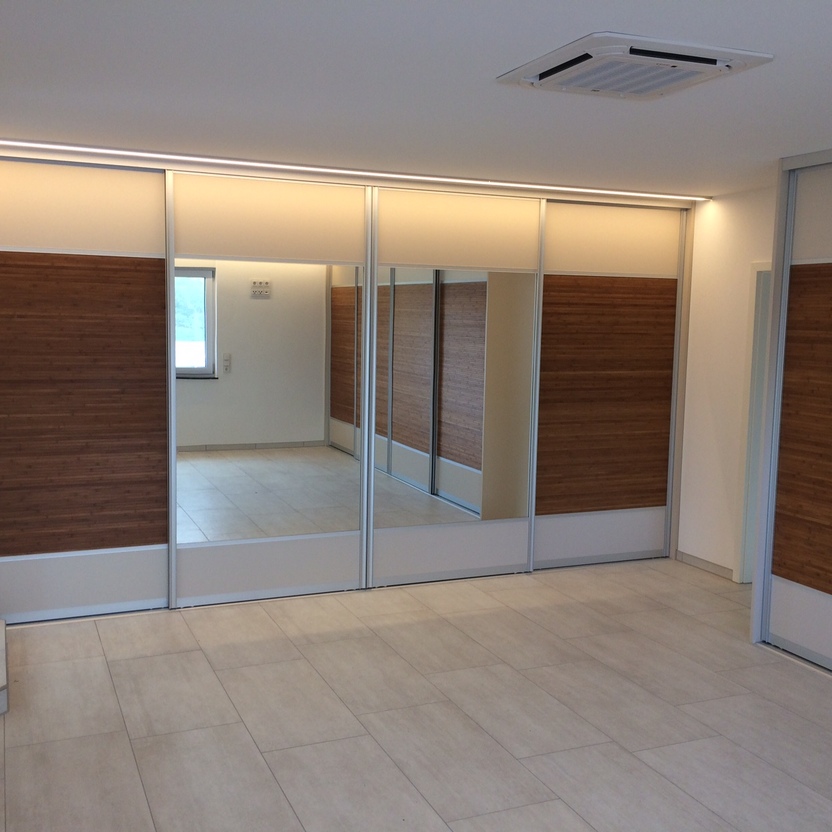 Großer Schiebetürenschrank mit Holzdekor und Spiegelfronten, beleuchtet, in einem hell gefliesten Raum mit moderner Decke.