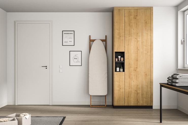 Ein hoher, maßgeschneiderter Holzschrank in natürlicher Maserung steht neben einem Bügelbrett, gegenüber einer weißen Tür und minimalistischer Wandkunst.